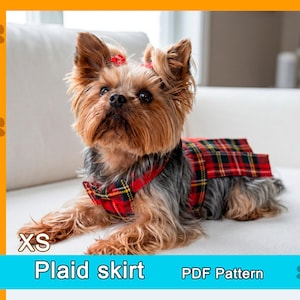 May include: A small dog wearing a red and black tartan skirt and matching collar. The dog has brown and black fur and is sitting on a white surface. Text on the image reads "XS" and "Plaid skirt".
