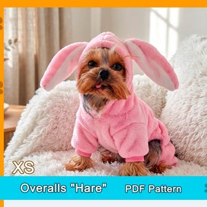 May include: A small dog wearing a pink bunny rabbit costume with long ears and a hood. The dog is sitting on a white, fluffy surface. The text "XS" is in the lower left corner, and "Overalls "Hare" PDF Pattern" is at the bottom.