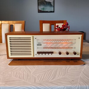 May include: A rectangular Wega radio with a wooden casing, a cream-coloured speaker grille on the left, and a glass-fronted control panel on the right. The panel features a glowing orange frequency display, five rectangular push buttons, and three circular knobs.