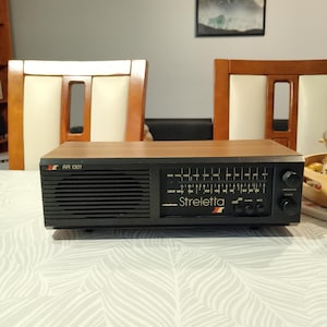 Op de afbeelding: Een rechthoekige tafelradio met een bovenkant in houtnerf en een zwart voorpaneel. De voorzijde is voorzien van een luidsprekerrooster aan de linkerkant, een frequentiedisplay met de tekst RR 1301 en Streletta, en zwarte knoppen.