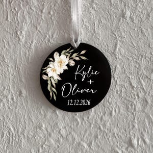 May include: A round black ornament featuring a white floral illustration on the left side. The text reads Kylie + Oliver with the date 12.12.2026 printed below in white script and sans-serif fonts. A white ribbon is attached to the top for hanging.