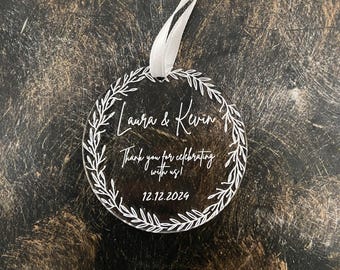 May include: A circular clear acrylic ornament featuring a white printed laurel wreath border. The text reads Laura & Kevin, Thank you for celebrating with us!, and 12.12.2024. A white ribbon is attached to the top for hanging purposes.