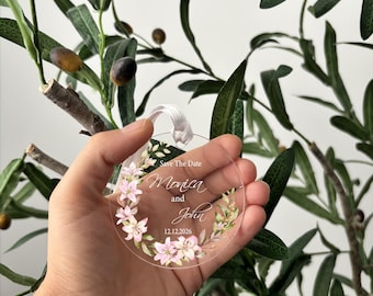 May include: A circular clear acrylic ornament featuring a printed floral wreath of pink lilies and green leaves. The text reads Save The Date, Monica and John, and 12.12.2026. A white ribbon loop is attached to the top of the transparent disc.