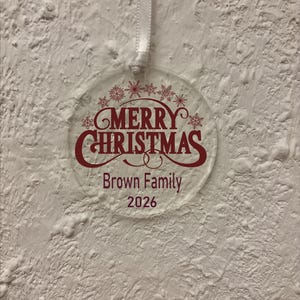 May include: A clear, circular acrylic ornament hanging from a white ribbon. The ornament features dark red text that reads Merry Christmas, Brown Family, and 2026, accented by a row of small, intricate snowflake designs above the text.