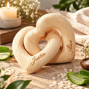 Può includere: Due cuori in legno di colore chiaro intrecciati con un motivo a venature naturali visibile. Il cuore a sinistra è inciso con il nome Justin in corsivo, mentre il cuore a destra è inciso con il nome Alice in un corsivo corrispondente.