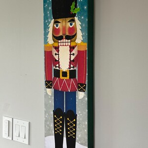 May include: A colorful illustration of a traditional nutcracker soldier with a red and white uniform, black hat, and a gold belt. The background is a blue and white snowy scene.