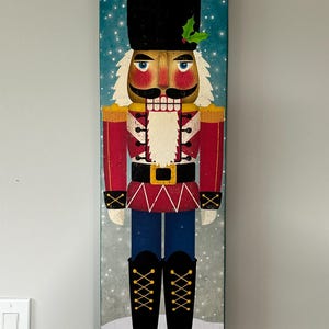 May include: A red and blue nutcracker with a black hat and a white beard. The nutcracker is standing on a snowy background with a blue sky.