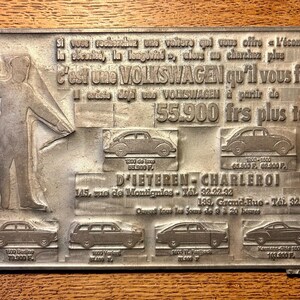 May include: A rectangular metal plaque featuring embossed text and illustrations of various Volkswagen car models. The text reads: Si vous recherchez une voiture qui vous offre l'économie, la sécurité, la longévité, alors ne cherchez plus C'est une VOLKSWAGEN qu'il vous faut.