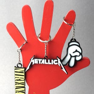 Könnte beinhalten: Drei Schlüsselanhänger mit schwarzen und weißen Logos auf einem roten handförmigen Display. Die Schlüsselanhänger zeigen die Bandnamen "Metallica", "Nirvana" und eine weiße Comic-Hand.