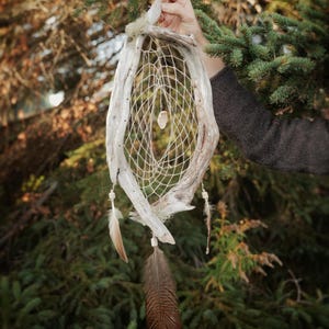 May include: A handcrafted dreamcatcher with a natural, rustic appearance. The oval frame is made from weathered wood, with a delicate web of woven string inside. Feathers and beads hang from the bottom, and a clear crystal is suspended in the centre.