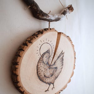 May include: A wood slice art piece featuring a hand-drawn illustration of a chicken with a human face and a halo. The artwork is suspended by twine from a curved wooden branch. The piece has a rustic, natural aesthetic.
