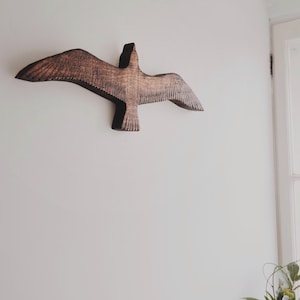 May include: A wooden wall art piece depicting a bird in flight. The sculpture is carved from dark brown wood, with detailed wing and feather textures. It is mounted on a white wall, creating a striking contrast. The bird's wings are outstretched, giving the impression of soaring.