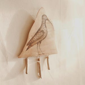 May include: A triangular wooden wall hanging featuring a detailed illustration of a bird with a long beak and speckled body. The bird is standing on a wooden base, with three cylindrical wooden accents hanging below. The artwork is in shades of brown and beige.