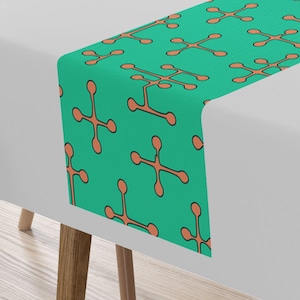 Könnte beinhalten: Ein türkisfarbener Tischläufer mit einem sich wiederholenden Muster aus orangefarbenen geometrischen Formen. Der Läufer liegt auf einer weißen Tischdecke mit einem Teller und Besteck auf dem Tisch.