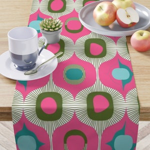 May include: A vibrant table runner with a retro-inspired design. The pattern features large, stylized floral shapes in pink, olive green, and teal, set against a white background. A cup, saucer, spoon, and apples are on the table.