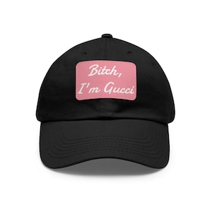 May include: A black baseball cap featuring a rectangular pink patch centered on the front. The patch displays the white text Bitch, I'm Gucci in a cursive script font. The cap has a curved brim and a structured crown with a button on top.
