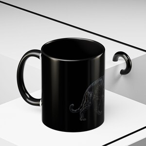 Peut inclure: Un mug en céramique noire orné d'une illustration subtile gris foncé représentant un grand félin au pelage tacheté. Le mug présente une finition lisse et brillante, un corps cylindrique et une anse incurvée sur le côté du récipient.