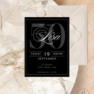 Op de afbeelding: Zwarte uitnodigingskaart met de tekst "Let's Toast Lisa" en het getal "50" in grijs. De uitnodiging bevat de datum, tijd en locatie details. Een witte envelop bevindt zich aan de rechterkant.