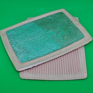 May include: Two rectangular, textured mats. One is light pink with vertical ridges, and the other is a light pink frame with a textured, green center. The mats are on a green background.