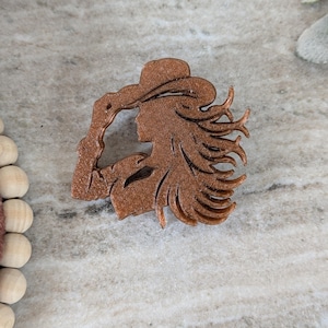 May include: A bronze-colored decorative cutout of a person wearing a cowboy hat, with flowing hair. The silhouette is detailed, showing the profile of the face and the hat's brim. The cutout is likely made of a solid material.