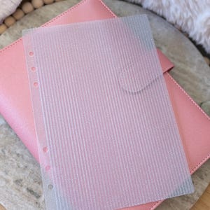 May include: A pink planner with a clear, textured, lined pocket sheet on top. The pocket sheet has three holes for binding. The planner is made of a smooth, pink material.