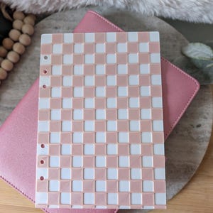 May include: A pink and white checkered planner insert with three pre-punched holes, resting on a pink planner cover. The insert has a woven appearance. The planner cover is on a wooden surface.