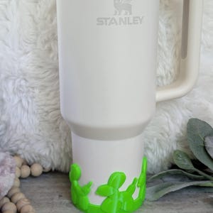 May include: A white Stanley tumbler with a handle and a silver rim. The Stanley logo is visible on the front. The bottom of the tumbler has a bright green decorative accent. The tumbler is sitting on a white, fluffy surface.