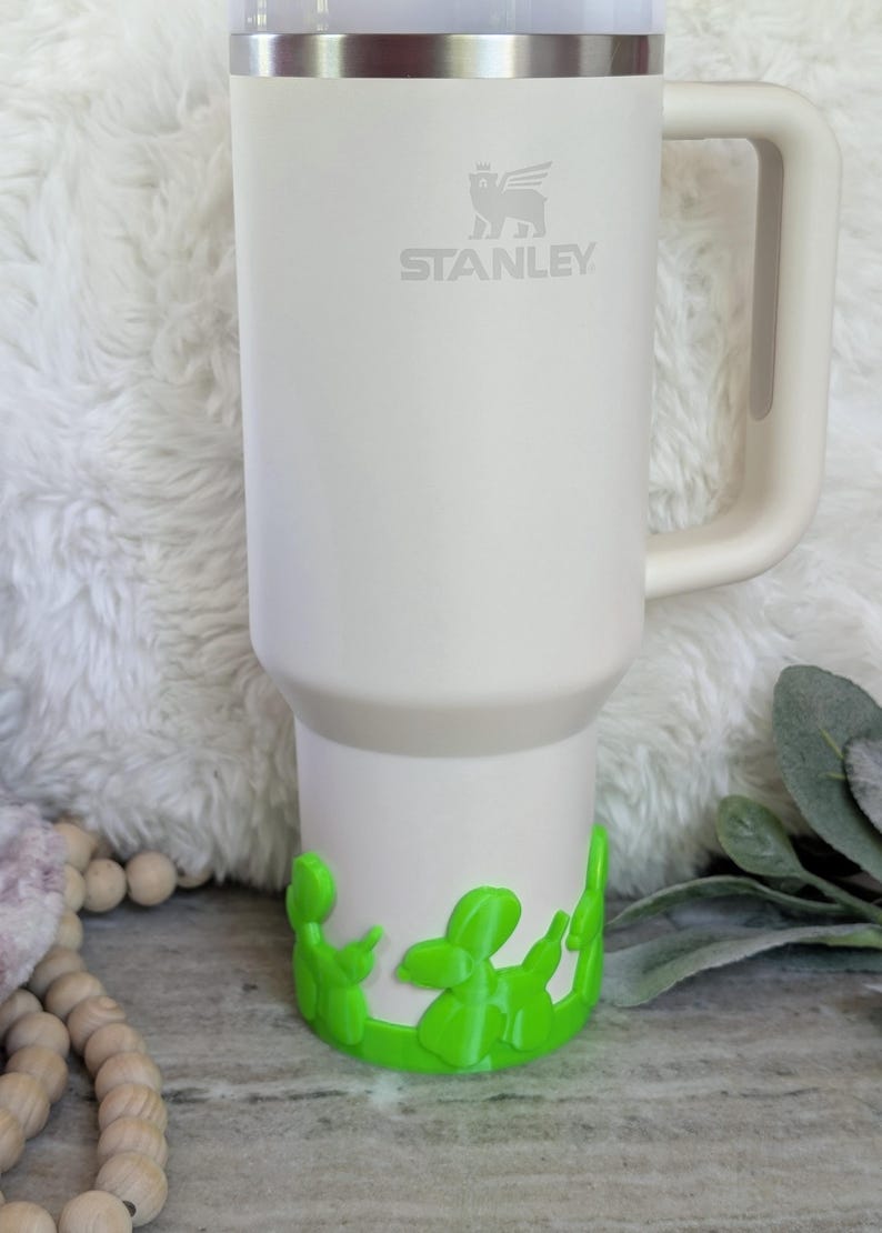 May include: A white Stanley tumbler with a handle and a silver rim. The Stanley logo is visible on the front. The bottom of the tumbler has a bright green decorative accent. The tumbler is sitting on a white, fluffy surface.