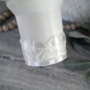 May include: A white tumbler with a decorative clear bow design around the base. The tumbler has a smooth, matte finish and a slightly tapered shape. The bow design adds a delicate, feminine touch to the tumbler.