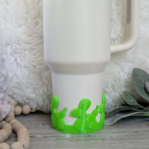 May include: A white Stanley tumbler with a green balloon dog base. The tumbler has a handle and the Stanley logo is visible. The balloon dog base is a bright green color and wraps around the bottom of the tumbler.
