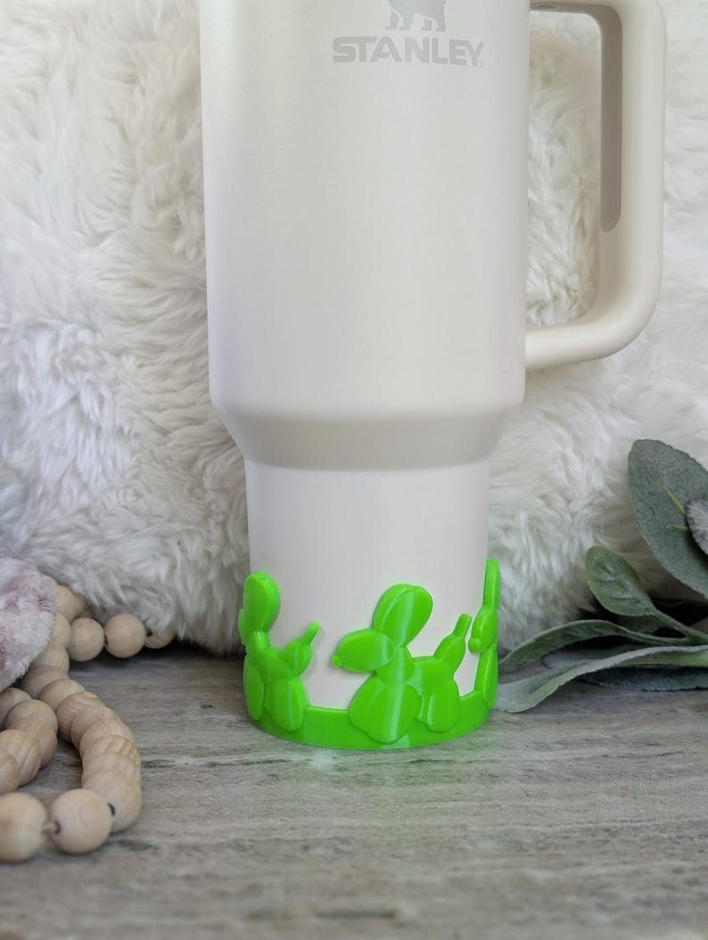 May include: A white Stanley tumbler with a green balloon dog base. The tumbler has a handle and the Stanley logo is visible. The balloon dog base is a bright green color and wraps around the bottom of the tumbler.