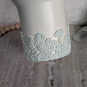 May include: A white tumbler with a light blue floral design around the base. The flowers are intricately cut out, creating a delicate and detailed pattern. The tumbler has a smooth, matte finish.
