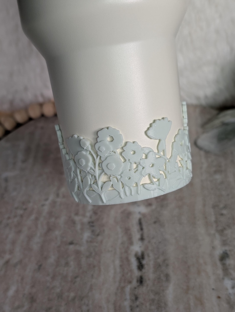 May include: A white tumbler with a light blue floral design around the base. The flowers are intricately cut out, creating a delicate and detailed pattern. The tumbler has a smooth, matte finish.