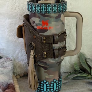May include: A camouflage-patterned Stanley tumbler with a brown leather holster featuring a tassel and turquoise accents. The tumbler has a handle and a straw. The Stanley logo is visible in orange.