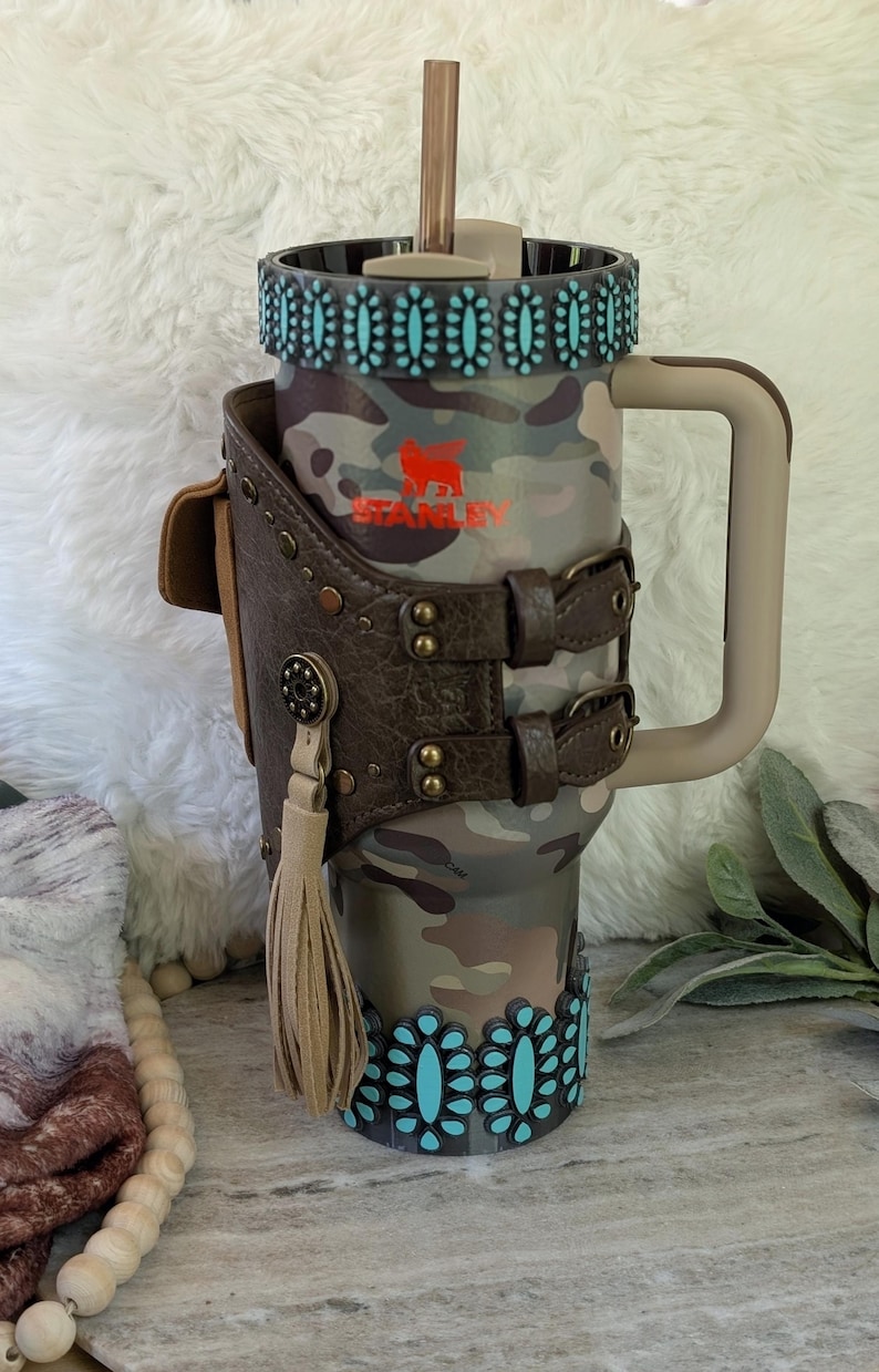 May include: A camouflage-patterned Stanley tumbler with a brown leather holster featuring a tassel and turquoise accents. The tumbler has a handle and a straw. The Stanley logo is visible in orange.