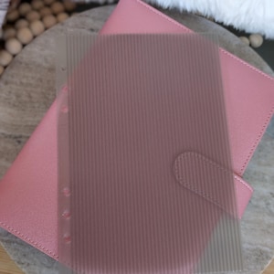 May include: A pink leather planner with a clear, vertically-lined plastic sheet overlay. The planner has a snap closure and is resting on a light-colored surface. The plastic sheet has three holes for binding.