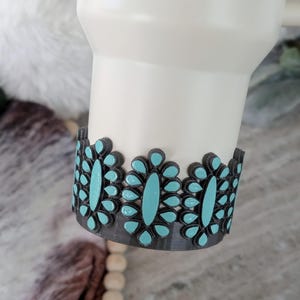 May include: A white tumbler with a decorative band featuring turquoise-colored oval accents set in a dark gray frame. The design wraps around the base of the tumbler, adding a pop of color and style.