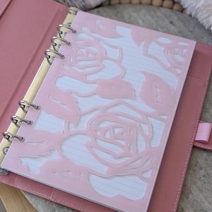 May include: Pink leather-bound planner with a rose-shaped cutout design on the cover. The planner is open, revealing lined paper. The planner has a gold-colored metal ring binder and a pink ribbon closure.