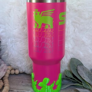 May include: A pink insulated tumbler with a green winged bear graphic and the number 5. The tumbler has a green cactus-shaped base and a blue lid with a green straw. The tumbler is designed to keep drinks cold or hot.