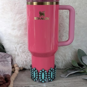 May include: A pink Stanley tumbler with a handle and a straw. The tumbler has a gold rim and the Stanley logo. A decorative black and turquoise floral design wraps around the base of the tumbler.