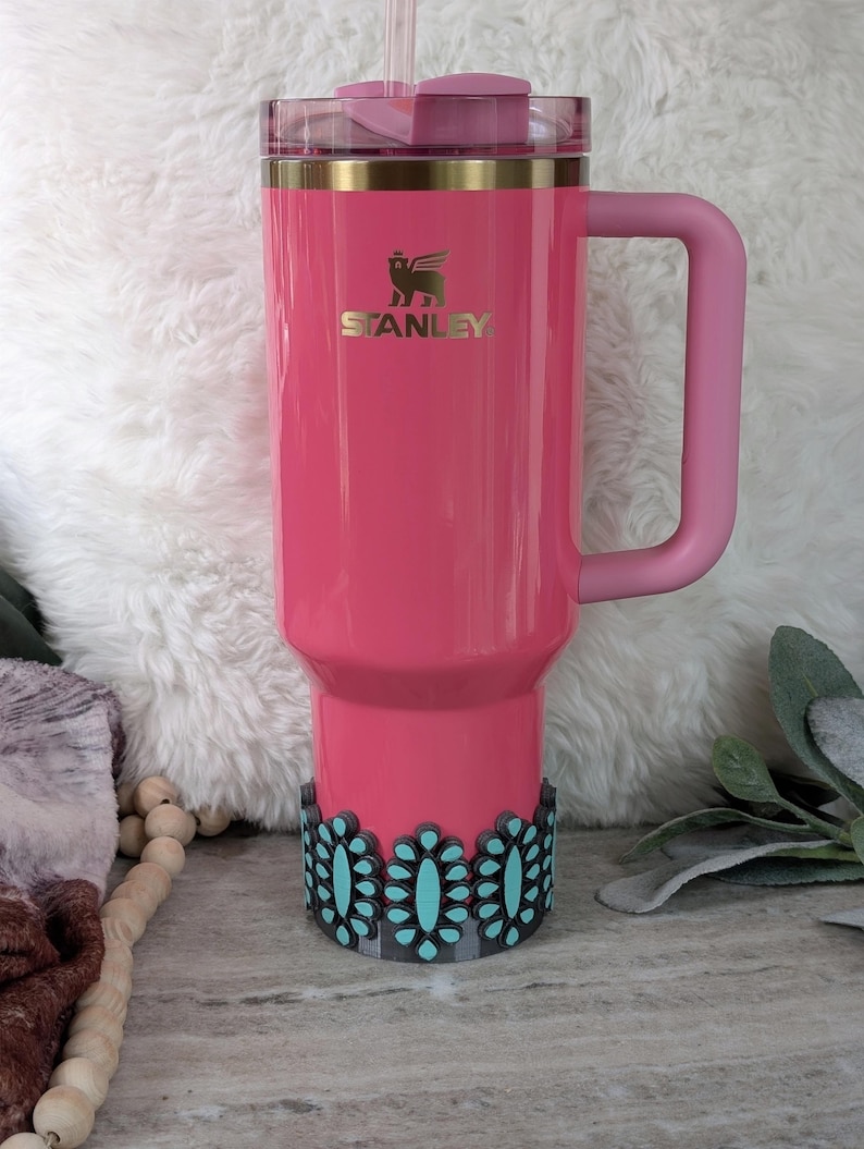 May include: A pink Stanley tumbler with a handle and a straw. The tumbler has a gold rim and the Stanley logo. A decorative black and turquoise floral design wraps around the base of the tumbler.