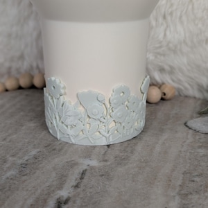 May include: A white ceramic mug with a floral design around the base. The flowers and leaves are in a light blue color, creating a subtle contrast against the white mug. The mug has a smooth, matte finish.