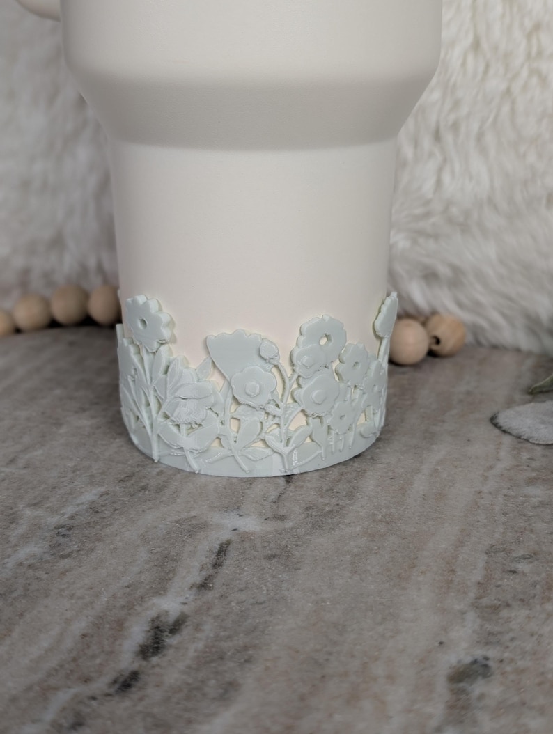 May include: A white ceramic mug with a floral design around the base. The flowers and leaves are in a light blue color, creating a subtle contrast against the white mug. The mug has a smooth, matte finish.
