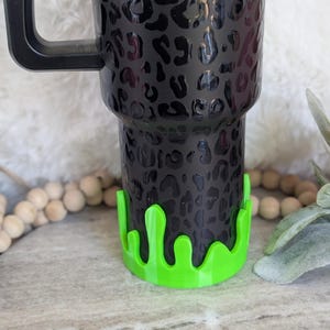 May include: Black tumbler with a leopard print design and a black handle. The base of the tumbler has a neon green drip design. The tumbler is designed to hold beverages.