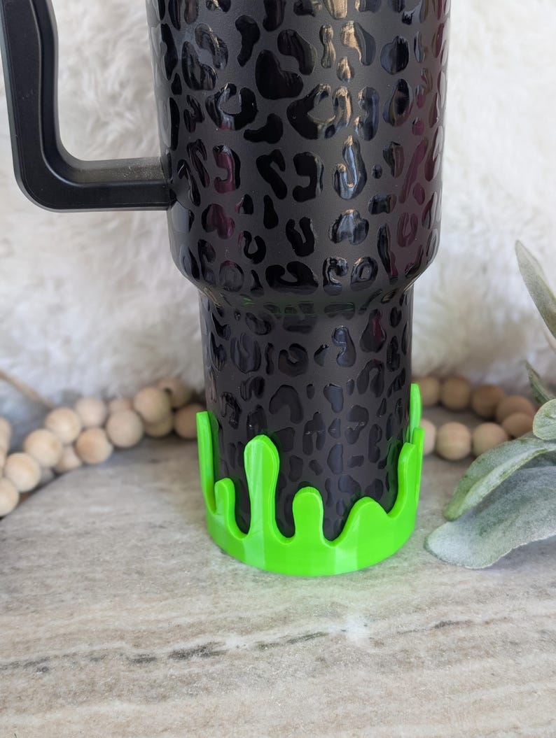 May include: Black tumbler with a leopard print design and a black handle. The base of the tumbler has a neon green drip design. The tumbler is designed to hold beverages.
