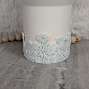 May include: A white tumbler with a floral design around the base. The flowers and leaves are intricately carved, adding a decorative touch to the tumbler. The tumbler is a solid white color.