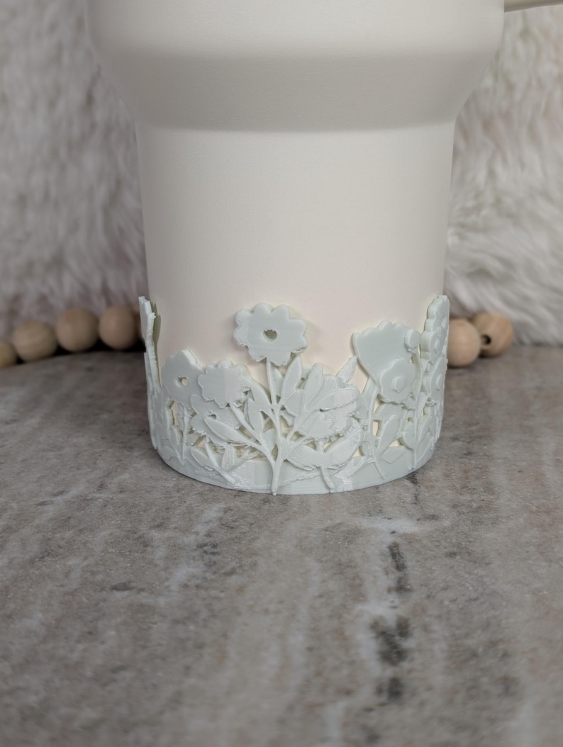 May include: A white tumbler with a floral design around the base. The flowers and leaves are intricately carved, adding a decorative touch to the tumbler. The tumbler is a solid white color.