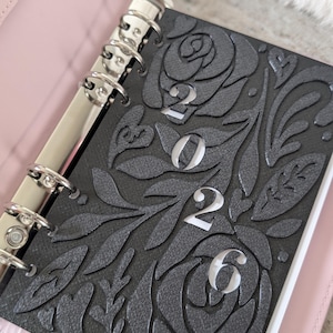 May include: A pink leather planner with silver rings, open to a black cover. The black cover features a floral design with the year "2023" cut out in white. The planner is open, revealing white pages.