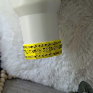 Puede incluir: Taza de viaje blanca con una banda amarilla que dice "CROSS CRIME SCENE DON'T CROSS". La taza tiene un asa y está colocada sobre un fondo blanco y esponjoso. La banda amarilla tiene diseños de flechas negras.