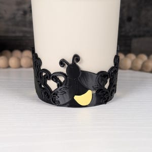 Puede incluir: Una banda decorativa negra que presenta la silueta de una luci&eacute;rnaga con un abdomen amarillo brillante, rodeada de intrincados patrones de hojas en filigrana. La banda est&aacute; colocada alrededor de un objeto cil&iacute;ndrico blanco liso, creando contraste.